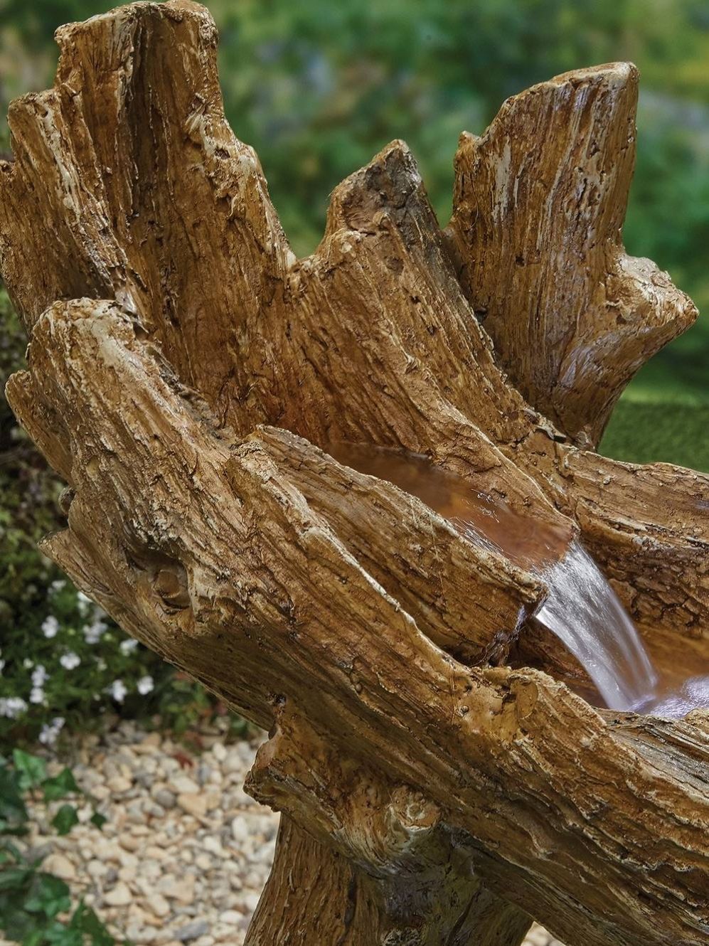 Introducing the Knotted Willow Falls with LEDs by La Hacienda, a realistic wooden log sculpture made from resin timber that serves as a water feature, with water softly flowing down its textured surface. The backdrop is blurred, highlighting lush greenery and delicate white flowers.