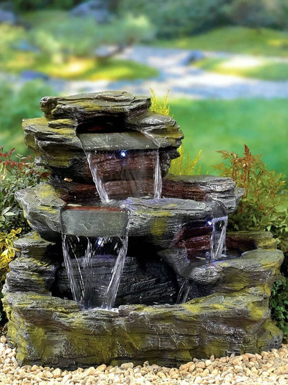 The Garda Falls inc LEDS by La Hacienda is a decorative tiered rock fountain that elegantly channels water down three levels. It is surrounded by small plants and pebbles, crafted from a durable resin structure. The built-in LED lights enhance its charm, set against the backdrop of a blurred, lush green garden.