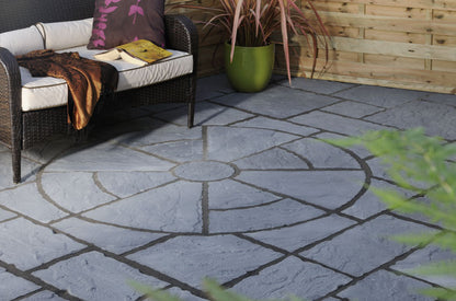 A cozy patio with a wicker sofa adorned with pillows, a throw blanket, and an open book. The floor features the Brisks Welsh Slate Rectory Circle Patio Kit by Bowland Stone, surrounded by rectangular stones. A green potted plant adds a touch of nature. Wooden panels line the perimeter, enhancing the garden landscaping charm.