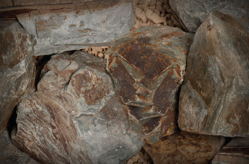 Close-up of Brisks' Rustic Slate Rockery by Bowland Stone, showcasing a pile of large, weathered rocks with diverse copper and grey hues. The rough textures and irregular shapes offer a natural, rugged look with small pebbles filling the gaps between the stones.