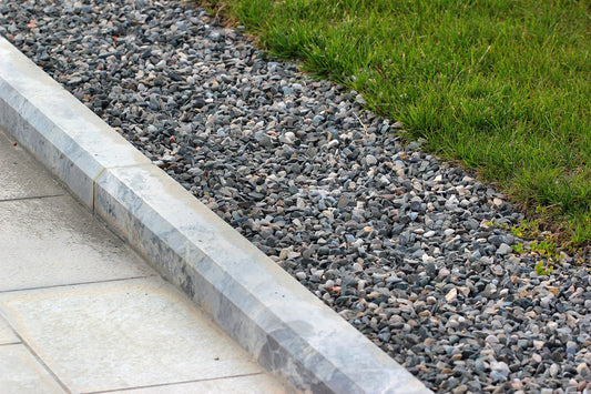 A close-up view of a landscaped edge, showcasing a section of green grass bordered by a strip of Brisks 14-20mm Silver Grey Granite Chippings, next to a paved walkway. The chippings create a clear separation between the grass and the pavement, enhancing both driveways and landscaping designs.