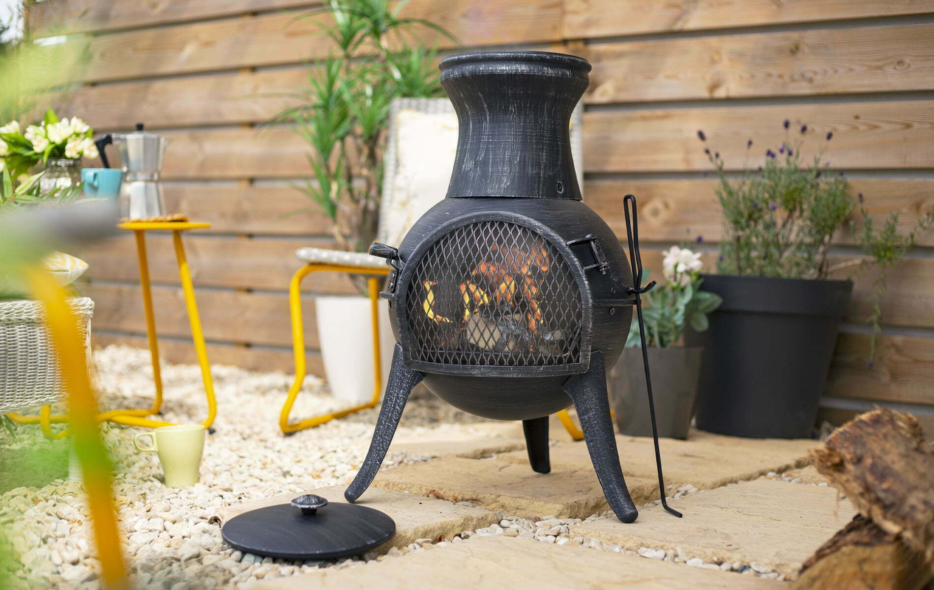 A La Hacienda Squat Chimenea, functioning as a patio heater, is situated on a beige stone patio encircled by white gravel. Positioned close to a wooden fence, it is accompanied by potted plants and a yellow table adorned with a coffee maker and cup. A black lid lies in front of the chimenea.