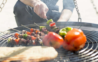 A person is grilling vegetable skewers and red bell peppers using the La Hacienda Tripod hanging Grill. A piece of flatbread sizzles nearby, with smoke rising as one hand adjusts a skewer. The sun shines brightly in the background, highlighting this perfect barbecue moment.