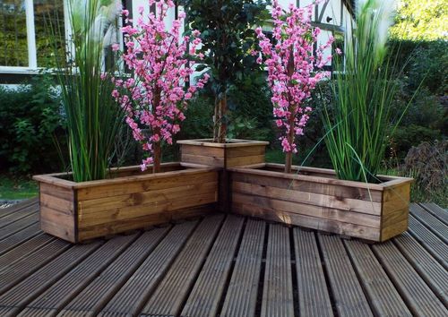 An English design wooden deck displays a Charles Taylor Trading Corner Planter Set with three rectangular planters made from sustainably sourced wood. It features lush greens, two pink flowering branches, and a central small tree, against verdant greenery and a building backdrop.