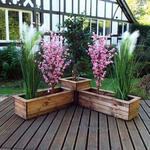 A patio corner displays the Charles Taylor Trading Corner Planter Set, crafted from sustainably sourced wood, with tall grasses and pink flowers. The planters form a zigzag on wooden decking, set against a charming half-timbered background.