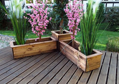 On the deck, a Corner Planter Set by Charles Taylor Trading, made from sustainably sourced wood, is filled with tall grasses and pink flowering plants in a triangular shape. An English-style garden area with shrubs is visible in the background.