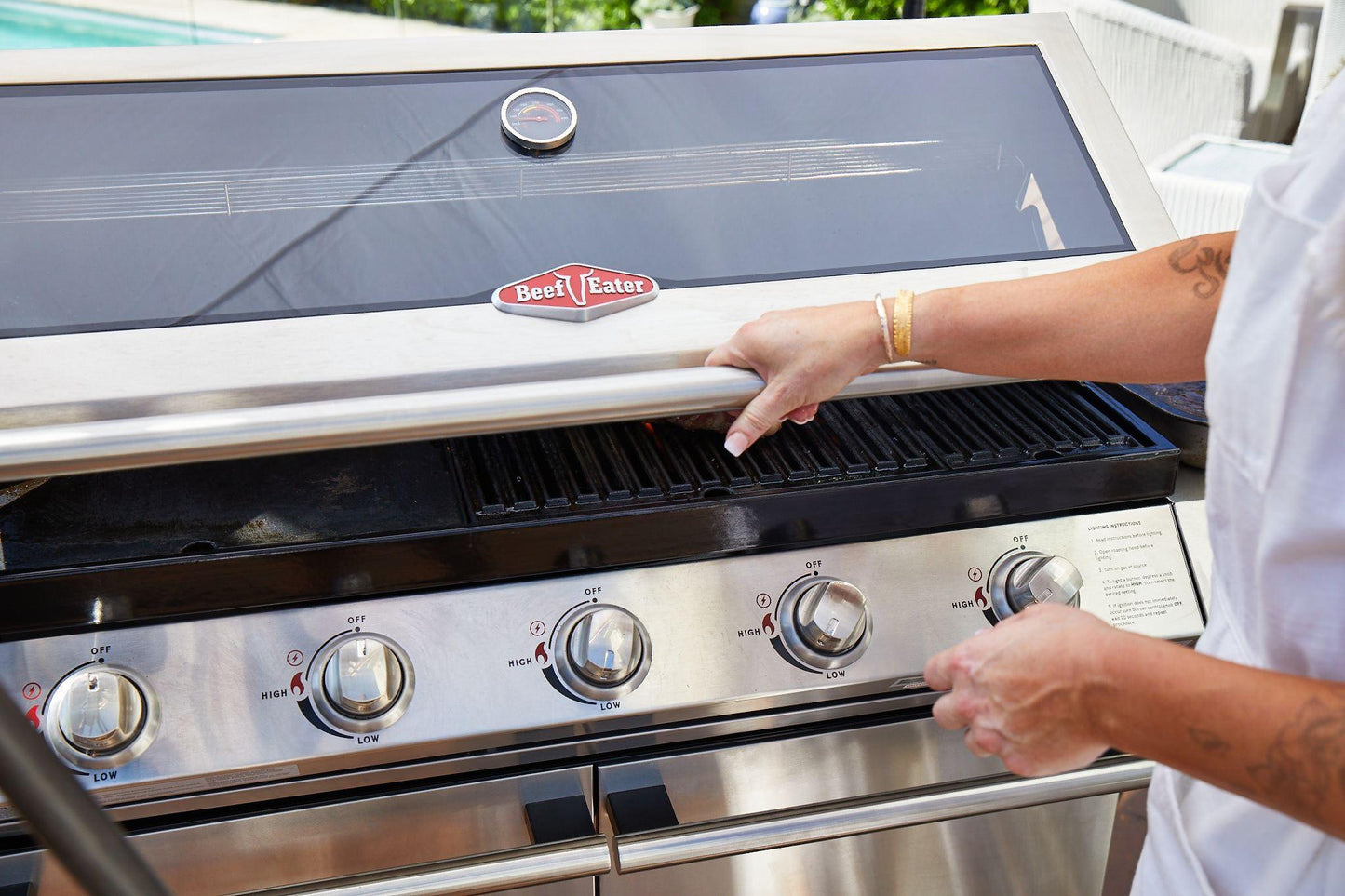 A person in a white shirt is opening the lid of a Brisks BeefEater 1200S Series 4 Burner BBQ & Trolley with four control knobs. The outdoor living setting is near a swimming pool with water steps in the background, exuding modern appeal. The person holds grilling tongs in one hand, ready for some BBQ action.