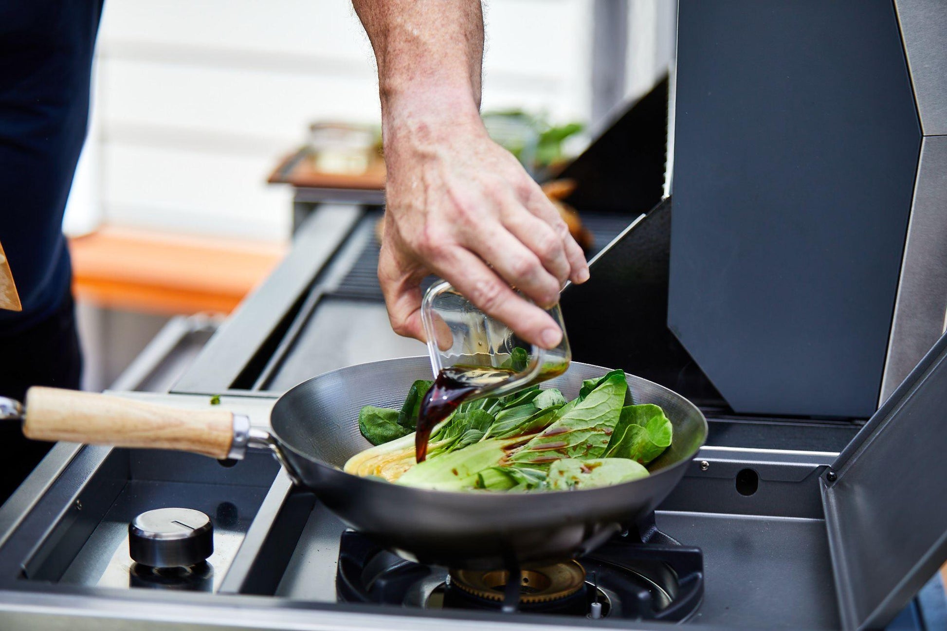 A person pours soy sauce from a small glass container into a wok filled with vegetables, including bok choy and green onions. The wok sits atop the sleek BeefEater 1500 Series 3 Burner BBQ & Trolley from Brisks, making for an outdoor cooking experience in what appears to be a modern kitchen.