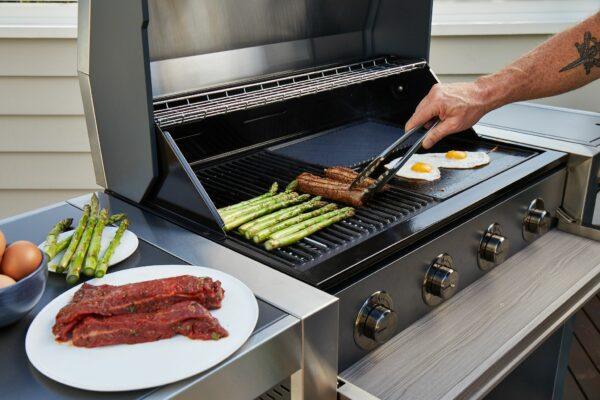A person grills vegetables, meat, and eggs on a Brisks BeefEater 1200E Series 5 Burner BBQ & Trolley. Asparagus, prime meat cuts, and a pair of eggs sizzle on the Brisks BeefEater 1200E Series 5 Burner BBQ & Trolley surface. Nearby, there's a plate of uncooked asparagus and seasoned meat waiting to enhance their outdoor living experience.