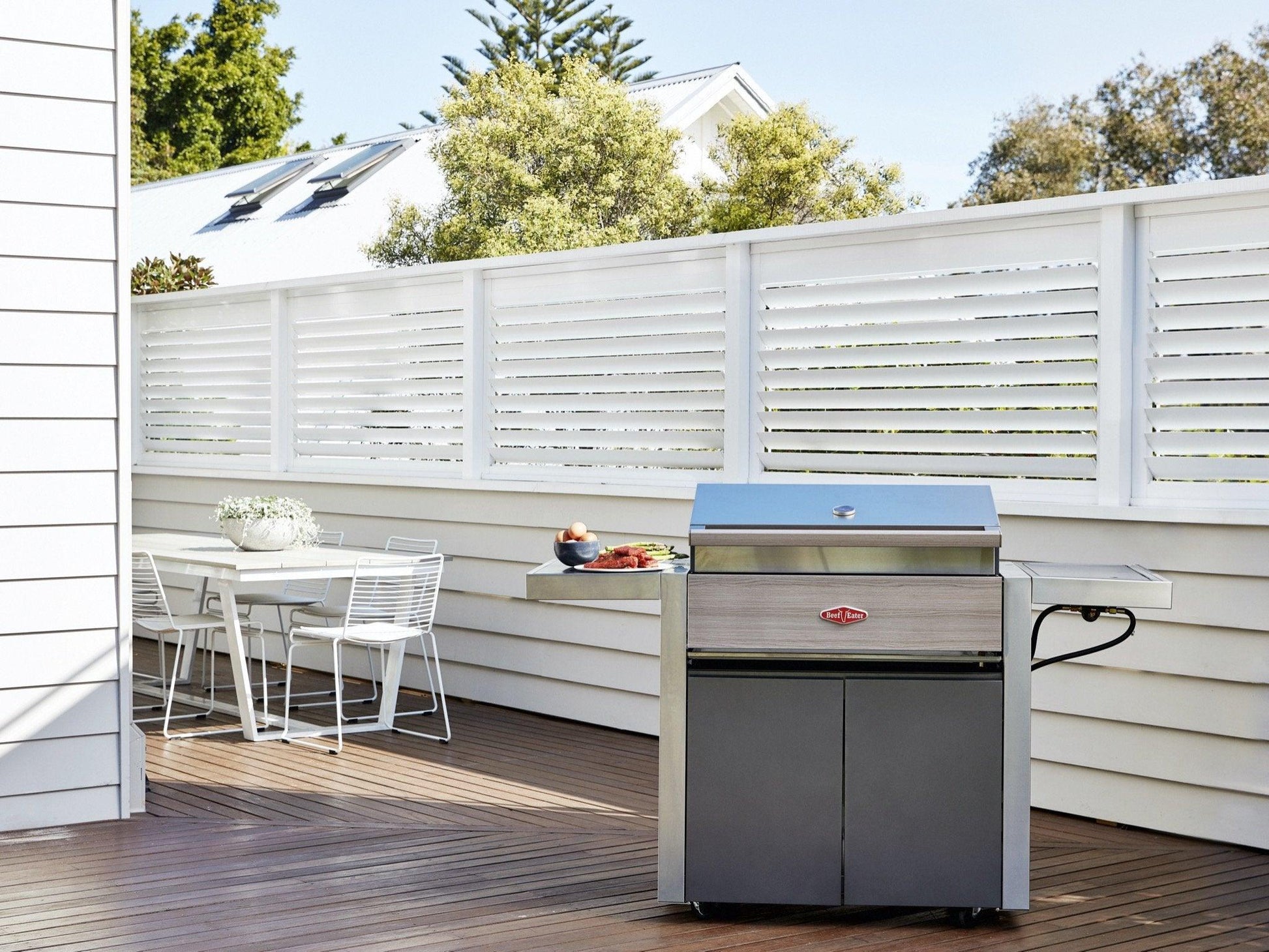 A modern outdoor patio features a Brisks BeefEater 1500 Series 3 Burner BBQ & Trolley on a wooden deck, next to a white dining table with three chairs. A potted plant adorns the table. The area is enclosed by a white privacy fence with horizontal slats. Trees and a house are visible in the background.