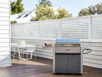 Outdoor patio with a closed Brisks BeefEater 1500 Series 3 Burner BBQ & Trolley, accompanied by a small table with four white chairs. The scene is set against a background of a white privacy fence and house, with some greenery visible over the fence, inviting you to an exceptional outdoor cooking experience.