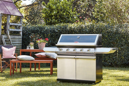 A shiny stainless steel Brisks BeefEater 1200S Series 5 Burner BBQ & Trolley stands on a neatly trimmed lawn. Next to it, a wooden table adorned with potted flowers and cushioned benches completes the perfect set-up for outdoor living in a sunny backyard surrounded by lush greenery and a wooded playhouse.
