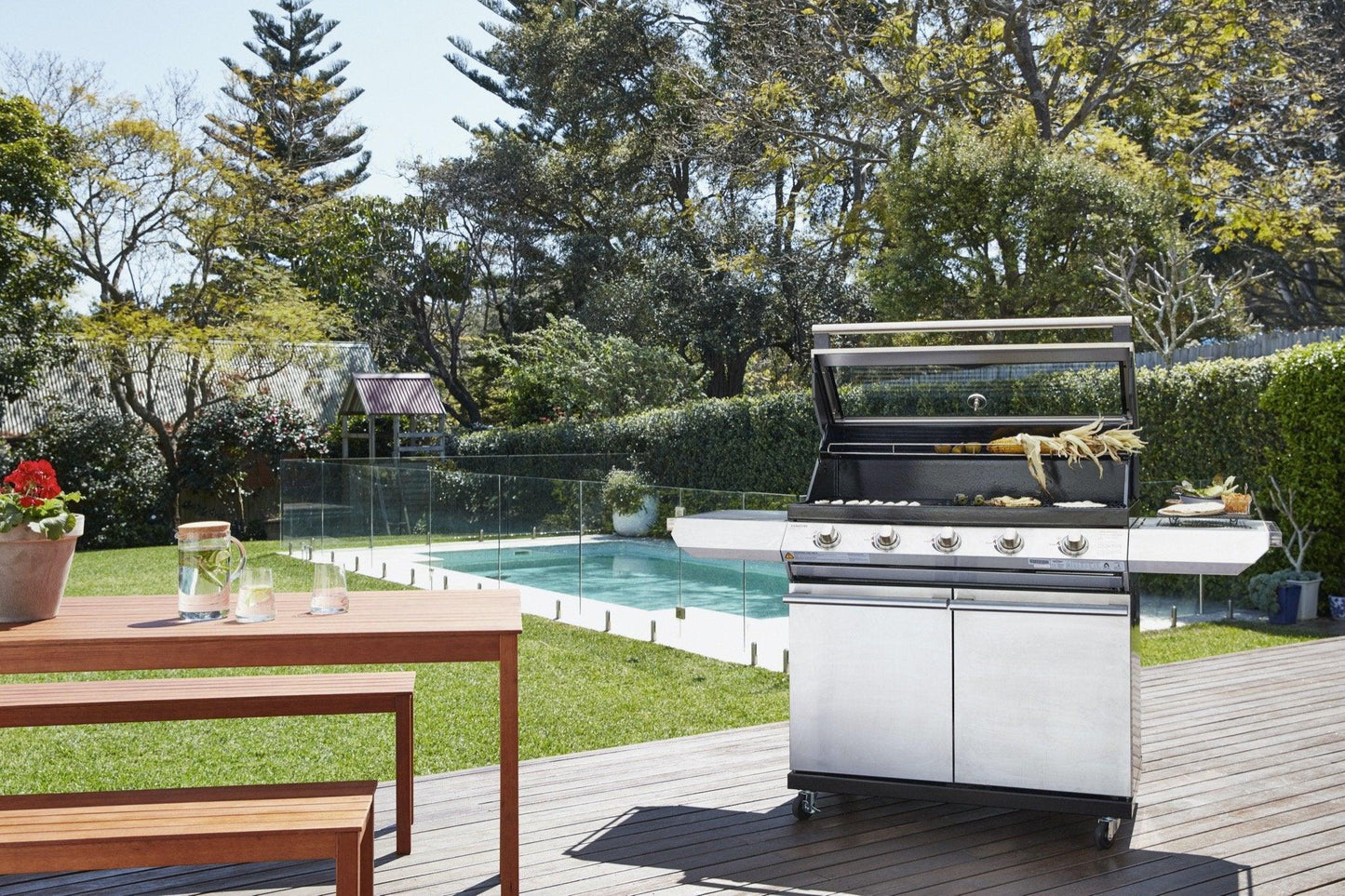 A Brisks BeefEater 1200S Series 5 Burner BBQ & Trolley stands on a wooden deck in a backyard, epitomizing outdoor living. The grill's lid is open, revealing food cooking inside. Nearby is a wooden table with a glass pitcher, glass, and a small plant. Behind the deck is a swimming pool enclosed by a glass fence. Trees surround the yard.