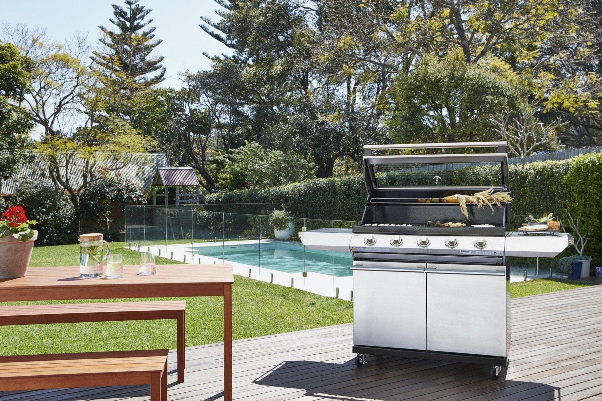 A modern outdoor patio showcasing a Brisks BeefEater 1200S Series 5 Burner BBQ & Trolley with its lid open and food grilling. The patio includes a wooden table with potted plants and drinks. A clear, fenced swimming pool is situated in a lush, green garden surrounded by trees and shrubs, perfect for outdoor living.