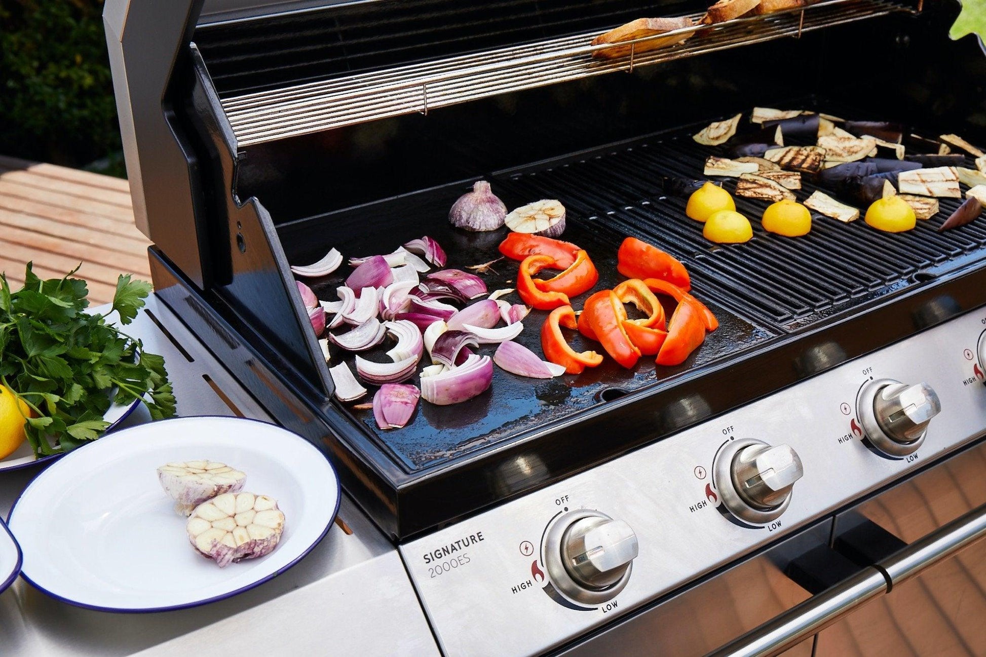 A Brisks BeefEater 1200E Series 5 Burner BBQ & Trolley with assorted vegetables cooking on it, including red onions, bell peppers, garlic, and yellow tomatoes. More vegetables await on a nearby plate and fresh herbs rest on a wooden surface, enhancing the outdoor living experience of this 5 Burner BBQ setup.