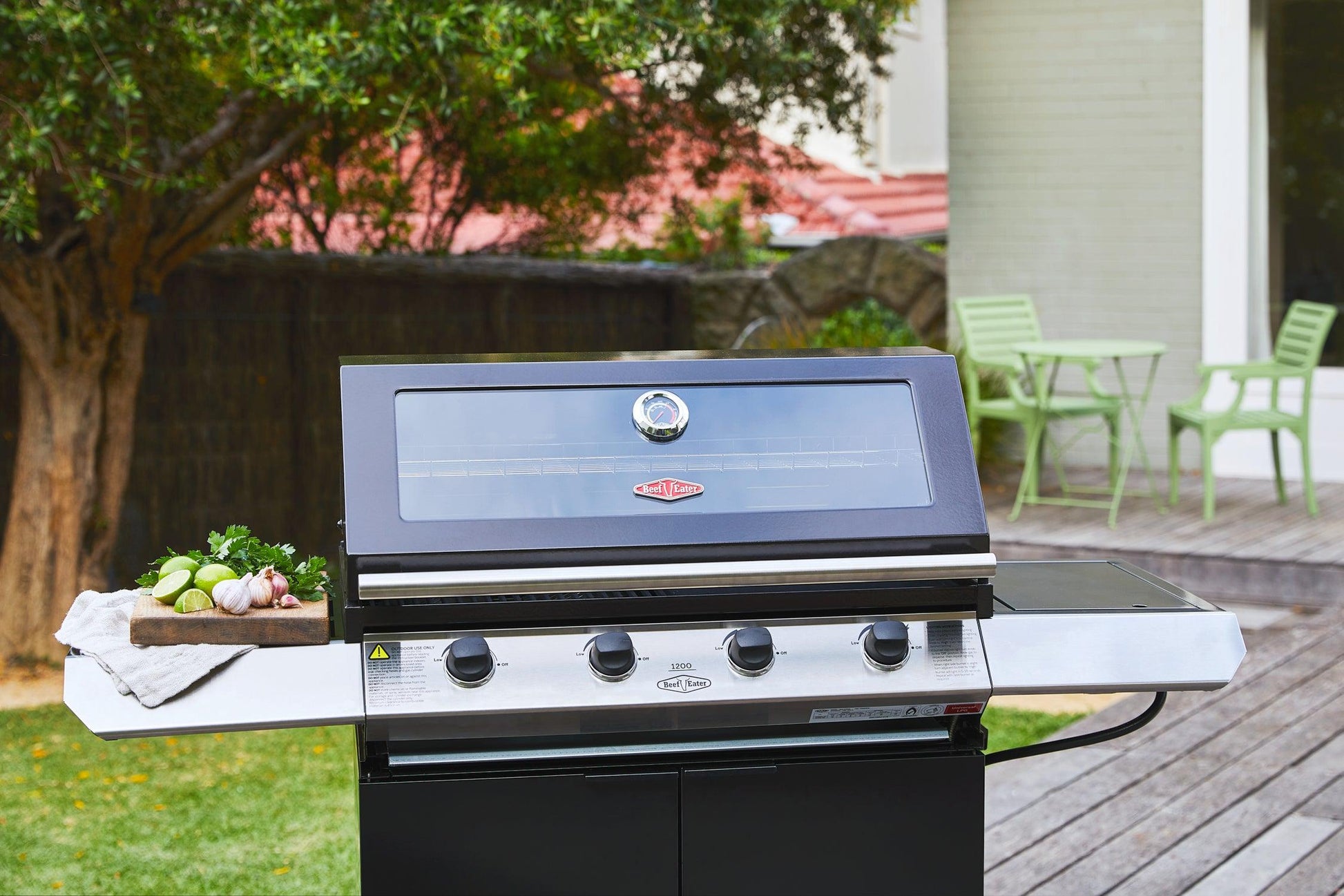 A black Brisks BeefEater 1200E Series 4 Burner BBQ & Trolley with four control knobs and a closed lid is situated on a wooden deck. A tray with vegetables and herbs adorns the left side shelf. In the background, an outdoor kitchen scene features a tree, a house, and a small seating area with a green table and chairs.