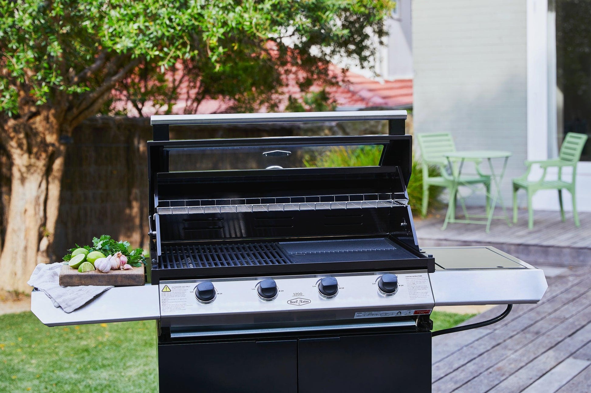 A Brisks BeefEater 1200E Series 4 Burner BBQ & Trolley stands proudly on a patio in a backyard with a tree and green chairs in the background. The grill has its lid open, revealing its potential for an outdoor kitchen, and a tray with vegetables and herbs is placed on its left side.
