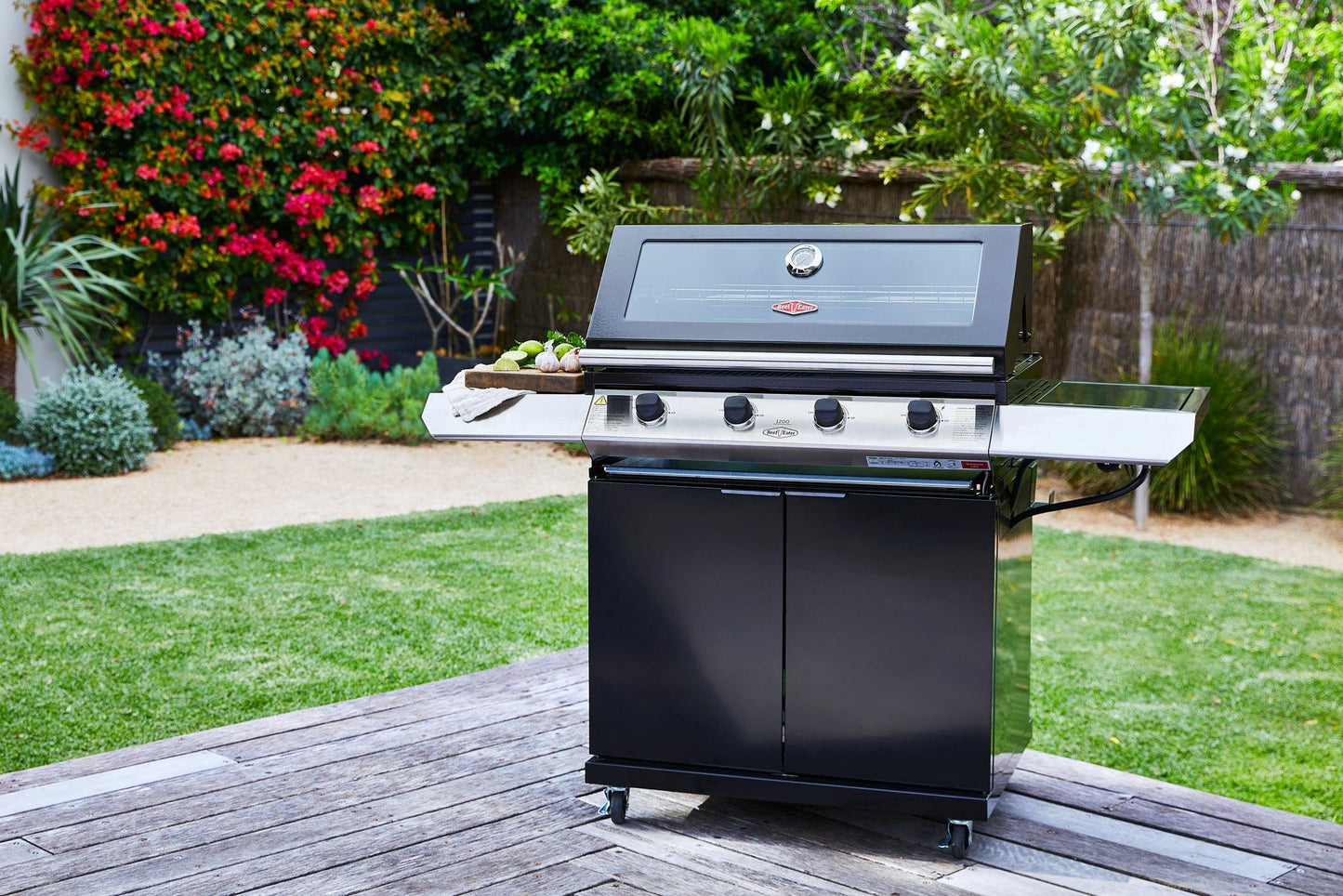 A black Brisks BeefEater 1200E Series 4 Burner BBQ & Trolley with four control knobs and a closed lid stands on a wooden deck in a garden. The grill has side handles and shelves, one holding vegetables. The background features green grass, various plants, bushes, and a tree with red flowers.