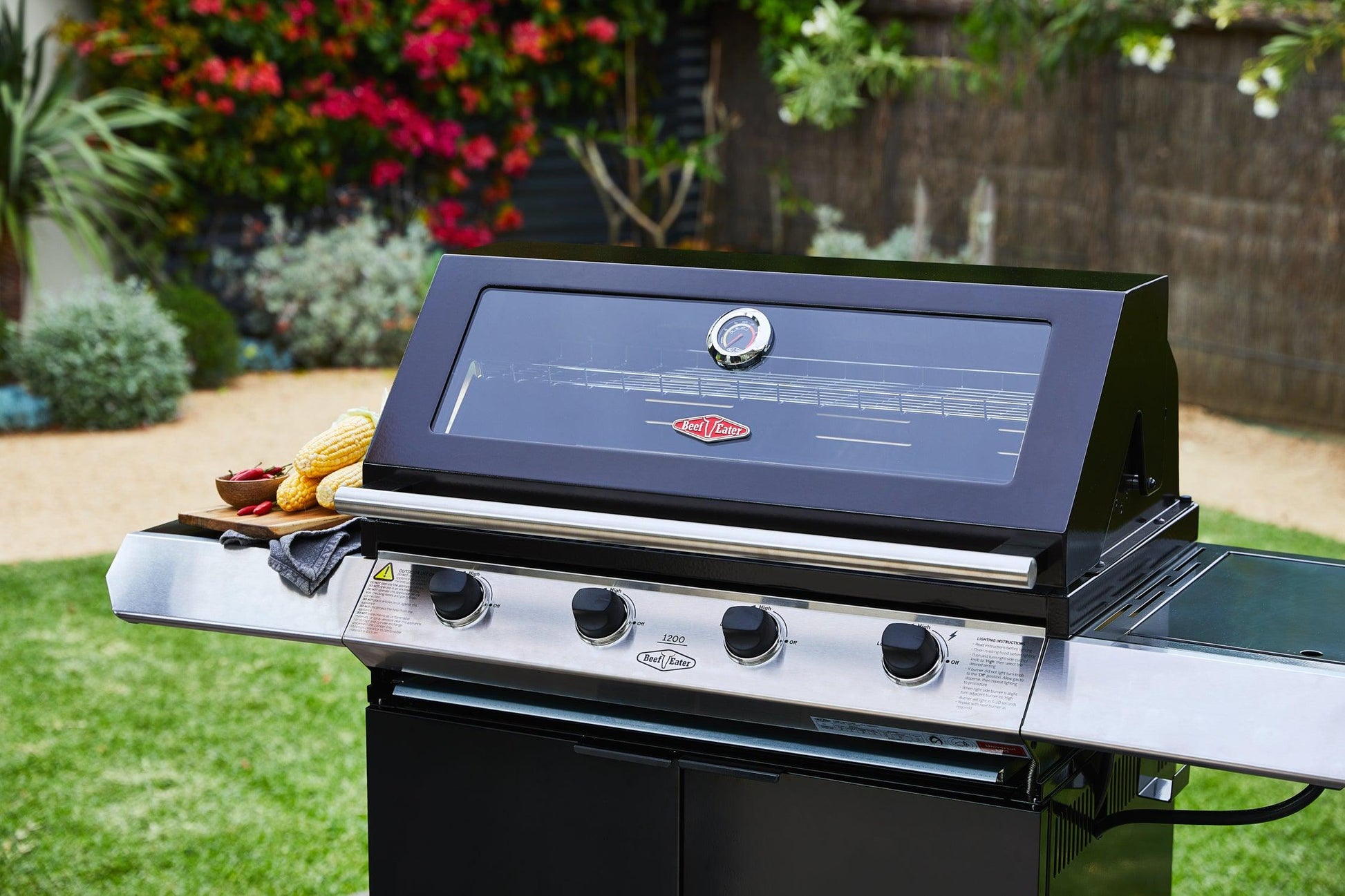 A Brisks BeefEater 1200E Series 4 Burner BBQ & Trolley stainless steel grill with four control knobs is placed on a garden patio. The grill, perfect for any outdoor kitchen, is topped with a glass viewing window, and corn on the cob and vegetables are visible on the side shelf. Colorful flowers and greenery decorate the background.