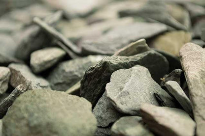 A close-up image of numerous grayish-green slate rocks, known as 20mm Green Slate Chippings by Brisks, piled together. The durable metamorphic rocks have a rough texture and irregular shapes, creating a natural and rugged appearance. Ideal for garden borders, the background is softly blurred to emphasize the details in the foreground.
