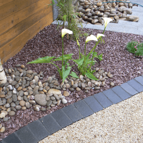 A garden bed with white calla lilies is bordered by large, smooth rocks. Dark brown mulch covers the bed, contrasting with black pavers. Pebbles fill the adjacent path that leads to a pile of rocks in the background. Brisks 20mm Plum Slate Chippings add a lilac hue to the scene, while a wooden structure is seen on the left.
