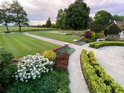 Beautifully landscaped garden with neatly cut grass, paved walkways of Kandla Grey Sandstone Paving Slabs by Brisks, and well-maintained flower beds featuring white, red, and green plants. Trees and a wooden fence can be seen in the background under a partly cloudy sky.