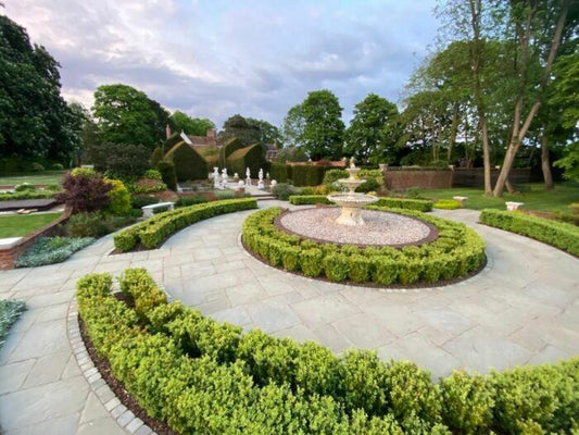 A serene garden featuring a circular stone pathway of Brisks Kandla Grey Sandstone Paving Slabs around a tiered fountain. Manicured hedges outline the path and surround the fountain area. In the background, statues, trees, and a neatly trimmed lawn rest under a partly cloudy sky with subtle grey hues.