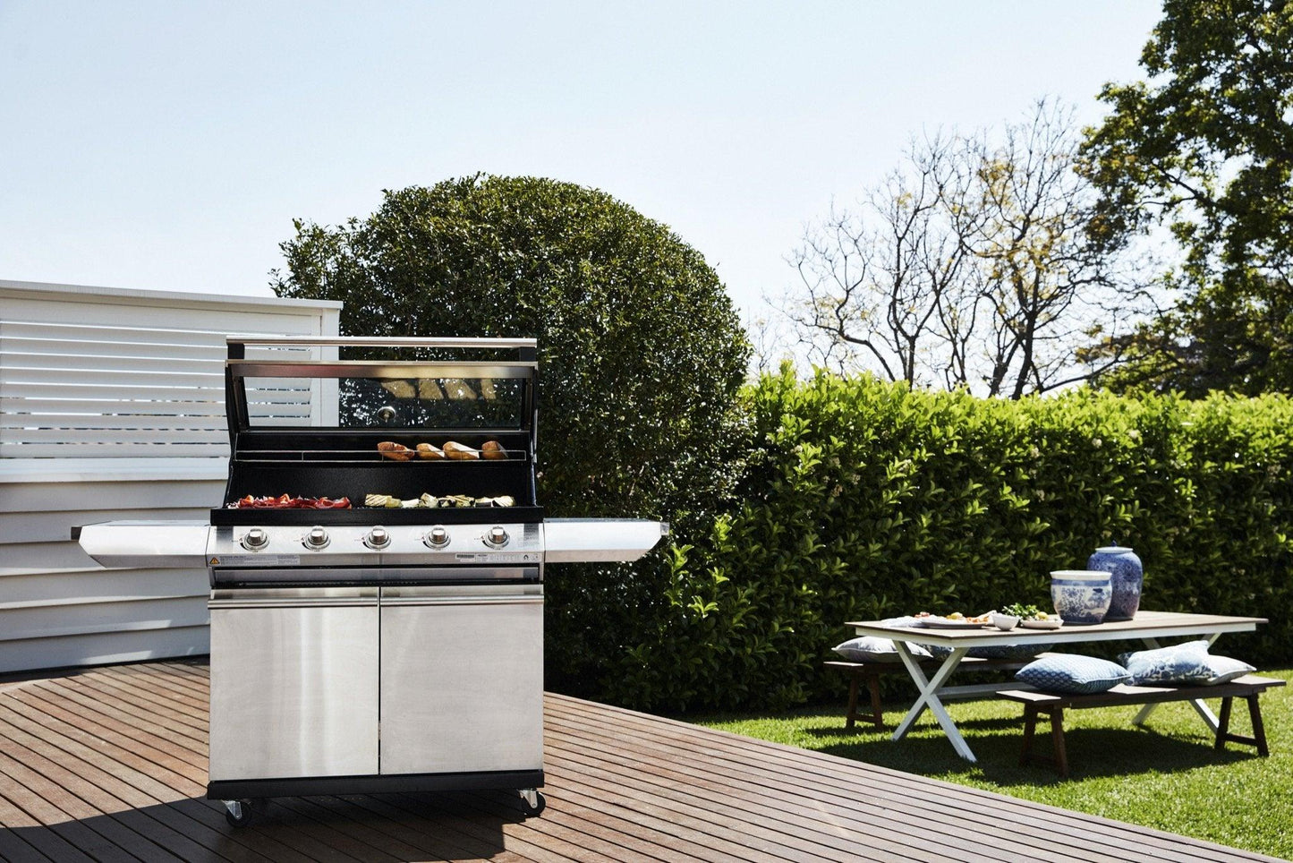 A sleek Brisks BeefEater 1200S Series 5 Burner BBQ & Trolley is positioned on a wooden deck, with various foods cooking on the stainless steel outdoor gas grill. In the background, there's a white wall and lush green hedges. Nearby, a white picnic table holds plates and vases, with cushioned seats on the grass, enhancing your outdoor living experience.