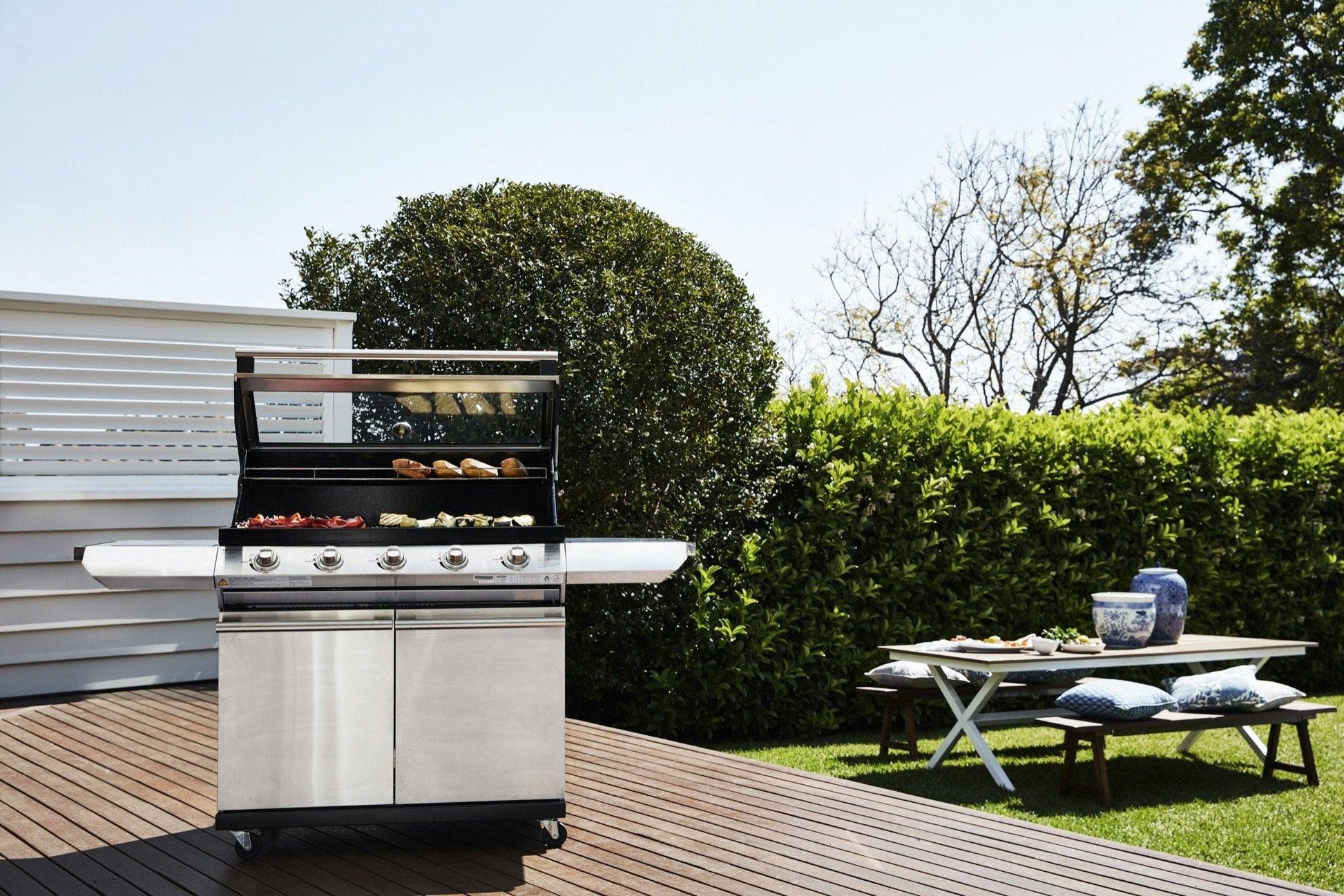 A Brisks BeefEater 1200S Series 4 Burner BBQ & Trolley with food items on the grates is situated on a wooden deck, embodying a modern appeal. Behind the grill is a lush hedge and clear sky. To the right, a low white table with a blue vase, plates, and cushions enhances the BBQ vibe on the grass.