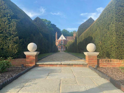 A brick pathway flanked by tall, neatly trimmed hedges leads to a small, picturesque brick house with a white, gabled roof. The entrance features two stone spheres on brick pillars and paving slabs crafted from Kandla Grey Sandstone Paving Slabs by Brisks. A blue sky with some clouds is visible in the background.