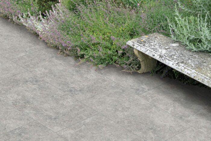 A stone bench with weathered texture is situated next to a garden bed filled with lush green foliage and small purple flowers. The ground beneath features Carmen Grey Porcelain Paving Slabs by Brisks, known for its slip-resistant surface and subtle silver hues. The scene appears tranquil and well-maintained.