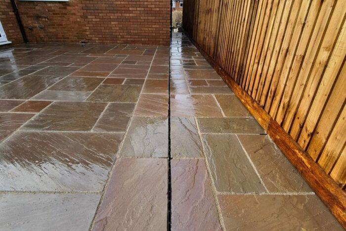A wet patio featuring large rectangular **Brisks Autumn Brown Sandstone Paving Slabs** in natural tones of brown and gray. The patio is bordered by a brick wall on the left and a wooden fence on the right. The Brisks sandstone paving slabs have a glossy finish from the rain.