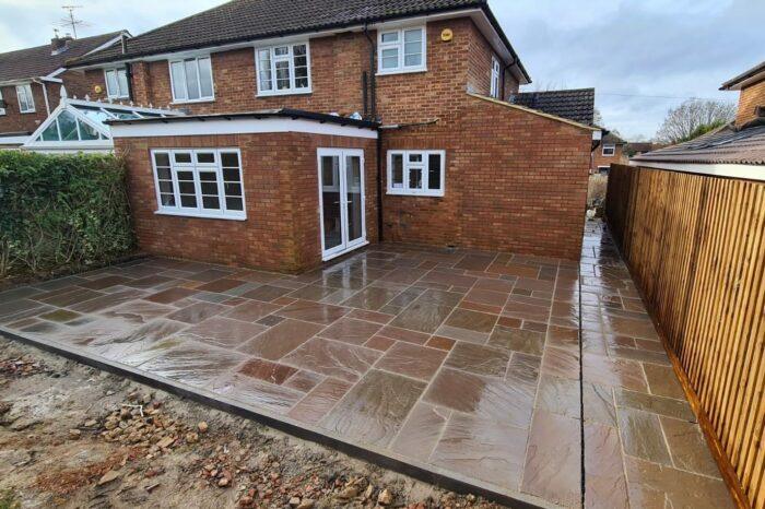 A recently built extension on a brick house with a fenced pathway on the right side. The paved backyard features large, wet Autumn Brown Sandstone Paving Slabs by Brisks, showcasing natural tones of brown and beige. A hedge borders the left side of the property. The sky is overcast.