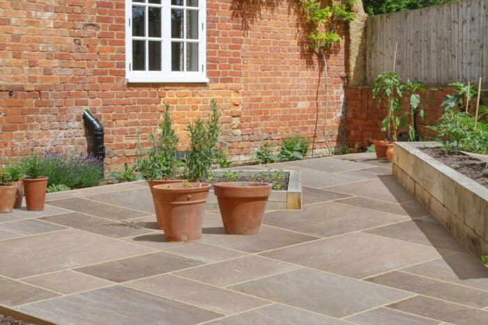 A backyard patio featuring large, Brisks Autumn Brown Sandstone Paving Slabs, redbrick walls, and a section of planter beds. Several potted plants in terracotta pots are scattered on the patio, while a patch of green foliage lines the base of the brick walls, complementing the natural tones.