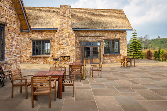 A scenic outdoor patio features rustic stone architecture with Brisks Autumn Brown Sandstone Paving Slabs, wooden tables and chairs, a large fireplace, and a neatly stacked woodpile. The area is surrounded by lush greenery and trees. The weather is clear with a partly cloudy sky.