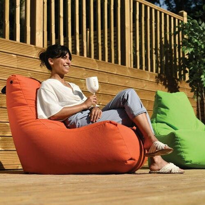 A person sits comfortably in an orange Brisks Extreme Lounging Outdoor Monster B-Bag, holding a glass and smiling. Another green Brisks Extreme Lounging Outdoor Monster B-Bag made from premium quality fabric is visible nearby. The background features wooden railings and greenery, highlighting the durable and comfortable setting.