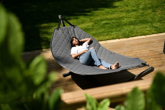 A person relaxes outdoors on a Brisks Extreme Lounging B-Hammock in a sunlit garden. They are lying on their back with one arm under their head, enjoying the sunny weather. The garden, featuring a lush green lawn and a wooden deck, complements the Brisks Extreme Lounging B-Hammock's durable and comfortable design for easy setup.