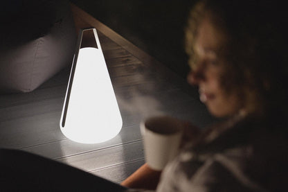 A person holds a steaming cup while sitting beside a triangular, cone-shaped Extreme Lounging B-Bulb Outdoor Light from Brisks that emits a soft white glow. The ambient lighting creates a cozy atmosphere, with the focus primarily on the outdoor light, perfect for any weatherproof adventures.