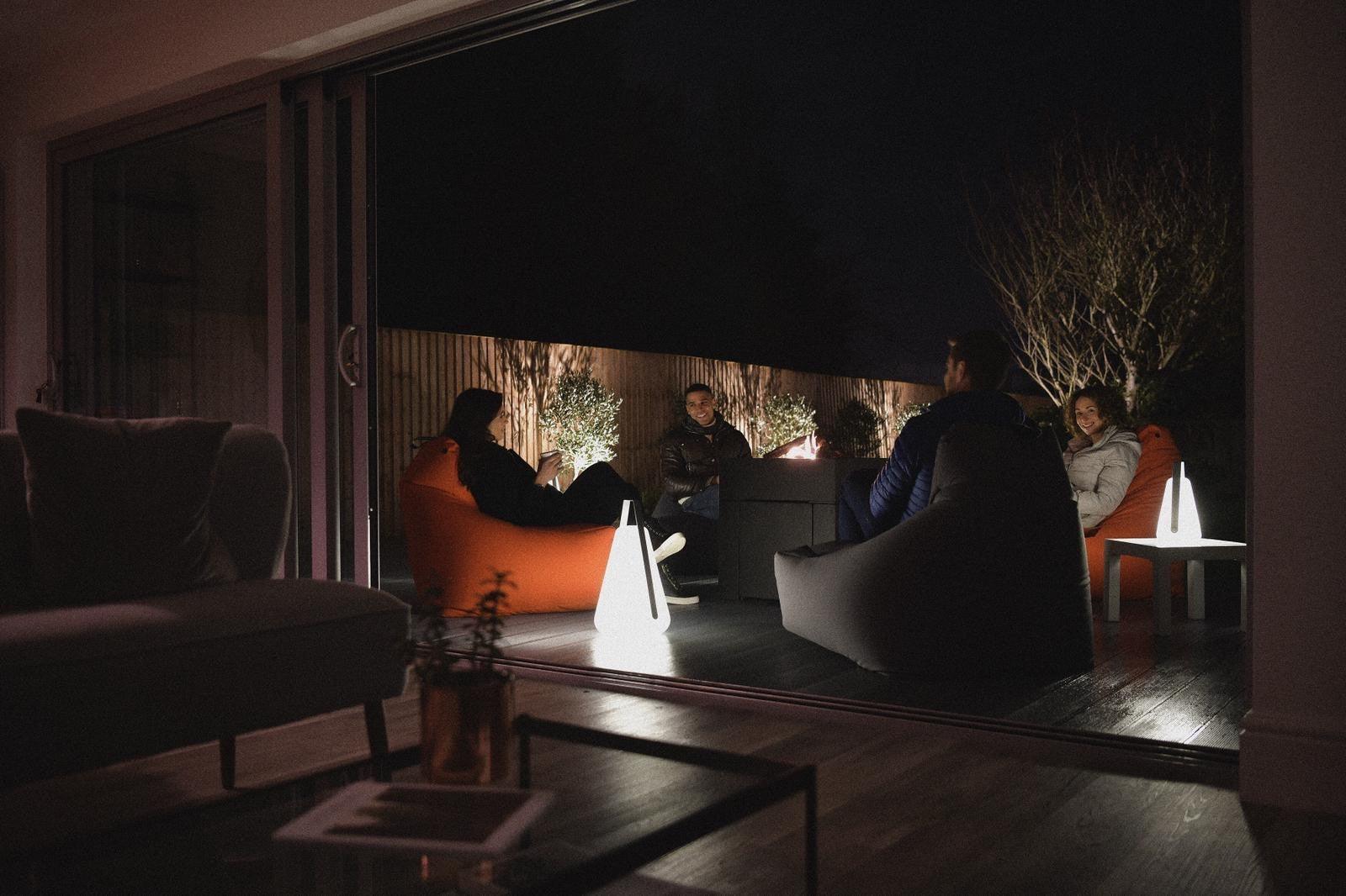A group of people sit on bean bags around a table on a deck at night. They are illuminated by Brisks Extreme Lounging B-Bulb Outdoor Lights and string lights along a wooden fence in the background. The setting appears cozy and intimate. Some indoor furniture is partially visible in the foreground.