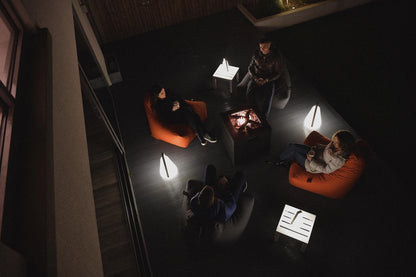 A bird's-eye view of five people sitting around a rectangular fire pit on an outdoor patio at night. They are seated on dark chairs with some additional light provided by modern, weatherproof, and rechargeable Brisks Extreme Lounging B-Bulb Outdoor Lights around them, creating a cozy and intimate atmosphere.