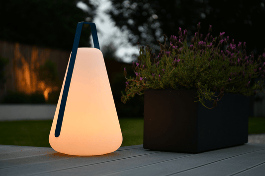 A glowing, cone-shaped Extreme Lounging B-Bulb Plus Outdoor Light from Brisks with a blue handle sits on a deck next to a rectangular black planter filled with purple flowers. The background shows a garden with trees and soft lighting, creating a serene evening atmosphere, perfect for this weatherproof outdoor light.