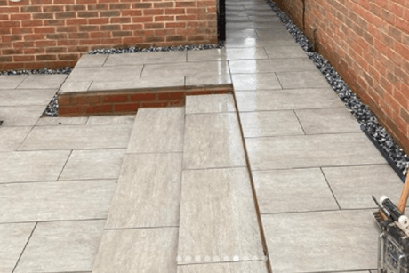 A paved walkway with large, light gray tiles runs alongside brick walls. The walkway has two narrow steps leading up to a higher, flat square area. 20mm Polar Black Ice Chippings by Brisks border the path, creating a neat and clean appearance.