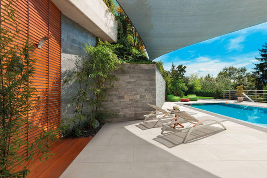 Modern outdoor patio area with two white lounge chairs beside a clear blue swimming pool. A gray stone wall, wooden door, and green plants add to the serene ambiance. The Brisks Frame Glen Porcelain Paving Slabs offer both style and durability, while non-slip paths ensure safety. Trees and a clear sky complete the scene.