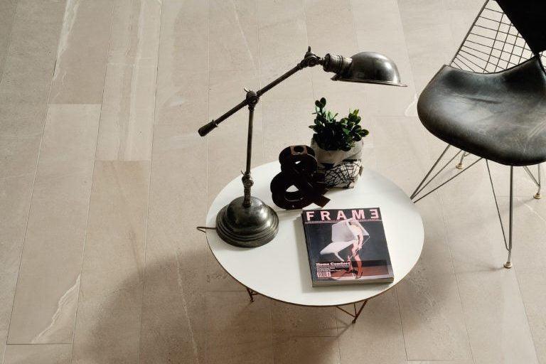 A white round coffee table with a metal desk lamp, a black ampersand symbol, a small potted plant, and a "FRAME" magazine on top sits gracefully. A wire-framed chair with a dark seat accompanies the setup on Brisks Britstone Sand Porcelain Paving Slabs.
