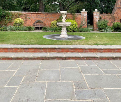 A stone fountain stands in the center of a manicured lawn, surrounded by shrubs and trees. Behind the fountain, there is a curved wooden bench against a brick wall with two arched openings. Stone steps, made of Brisks Kandla Grey Sandstone Paving Slabs, lead from a patio area up to the lawn.