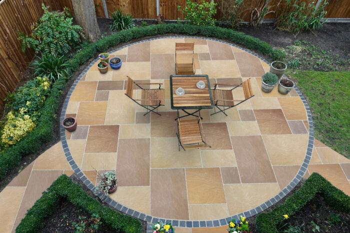 A circular patio with beige and brown tiles is centered in a garden. Featuring a four-piece wooden table set and surrounded by plants, potted flowers, and bordered by a low hedge, the Sunset Buff Sandstone Paving Slabs by Brisks add charm. The patio is elegantly encircled by dark brick edging.