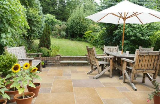 A sunny patio features wooden furniture, including a dining table, chairs, and a bench, all arranged on Brisks Sunset Buff Sandstone Paving Slabs. Large potted sunflowers decorate the space. An umbrella provides shade, and lush green vegetation surrounds the area, extending to a grassy lawn.