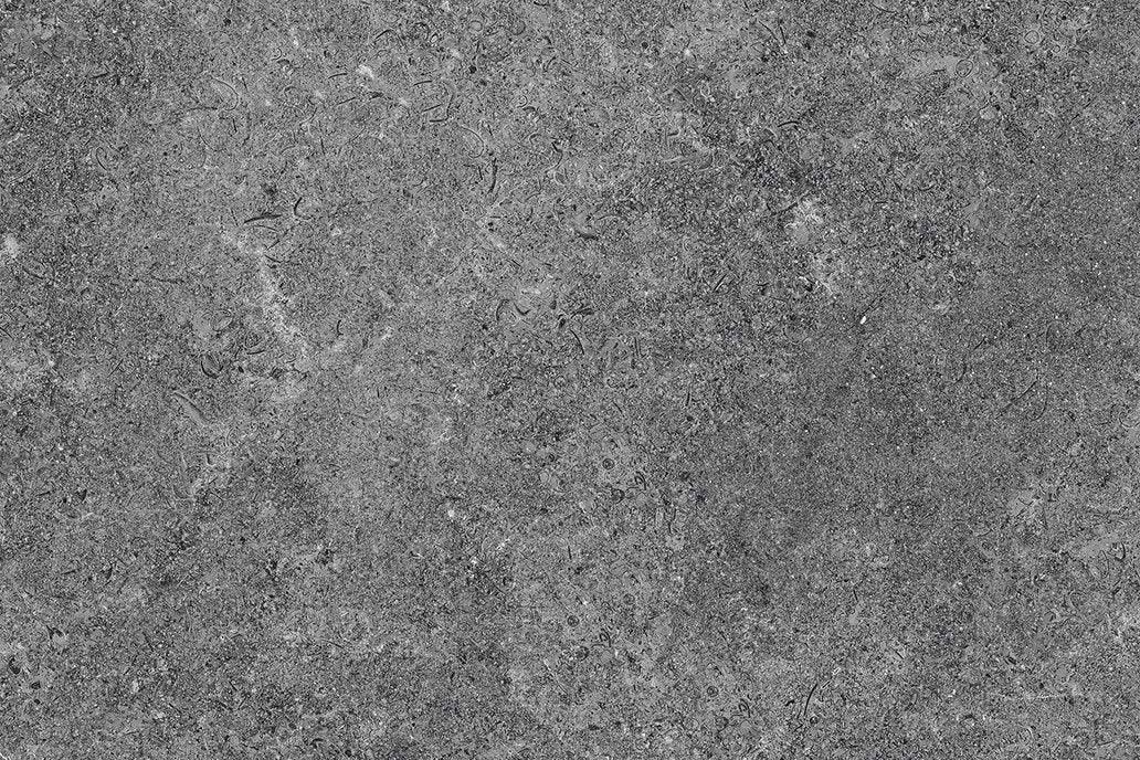 Gray concrete surface with a rough, speckled texture, featuring small pebbles and stones embedded within it. Perfect for landscaping projects, the Carmen Anthracite Porcelain Paving Slabs by Brisks show subtle variations in color from light to dark gray, and some faint, irregular lines and patterns presumably caused by wear or weathering.