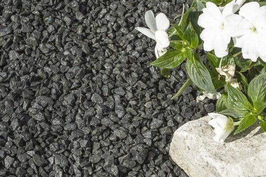 A close-up of black basalt gravel ground cover with a white flowering plant and a piece of light-colored stone in the corner. The white flowers and green leaves create a striking contrast against the Brisks 10-20mm Black Basalt Chippings background.