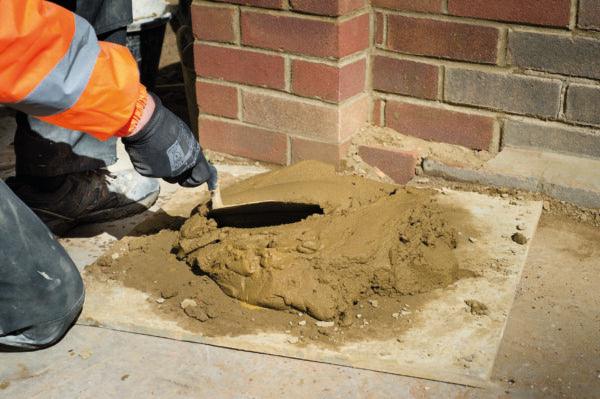 A construction worker, wearing a high-visibility jacket and gloves, is seen kneeling on the ground, using a trowel to smooth out Brisks Mortar Mix Concrete on a flat board. The worker is next to a brick wall corner, expertly performing bricklaying tasks.